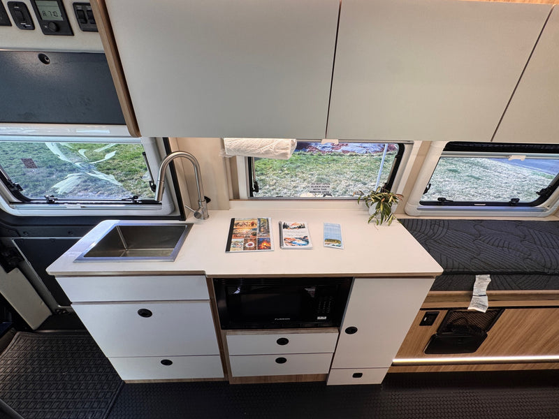 Inside an RV rental: a compact kitchen with sink, Furrion microwave, and storage, adjacent to a sleeping area. Bright windows reveal a grassy exterior. — photo 15