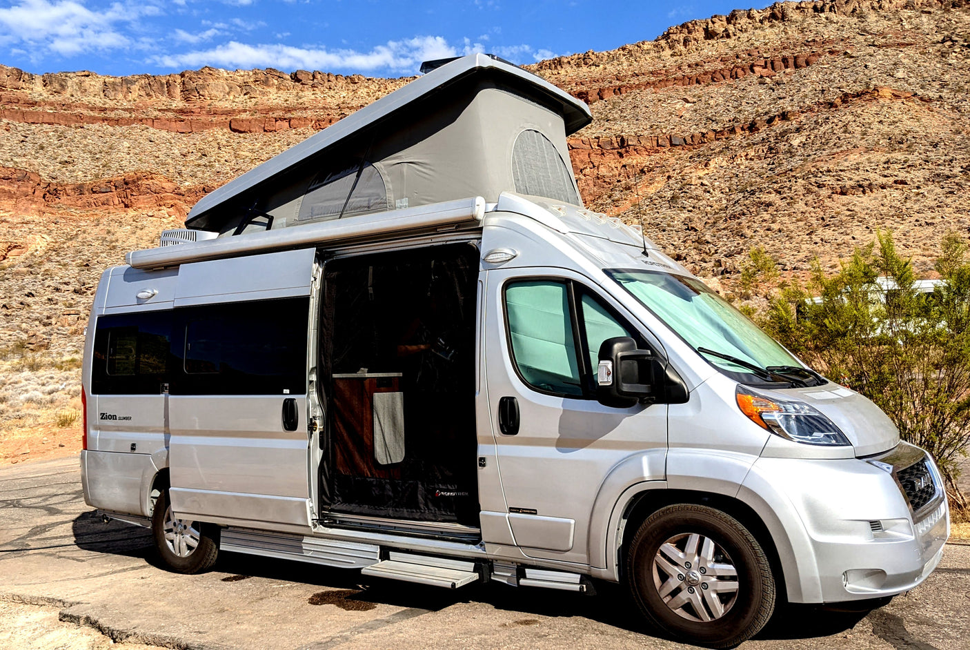 Silver pop-top camper van (RV rental) parked on asphalt, revealing its open screen door against rocky desert mountains.