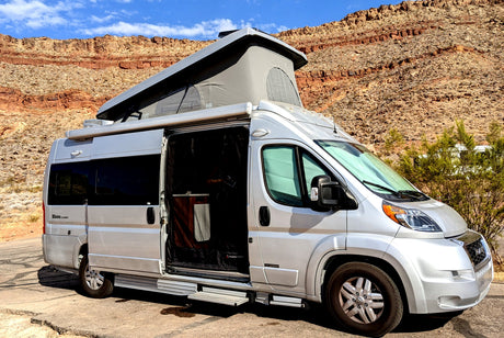 Silver pop-top camper van (RV rental) parked on asphalt, revealing its open screen door against rocky desert mountains.