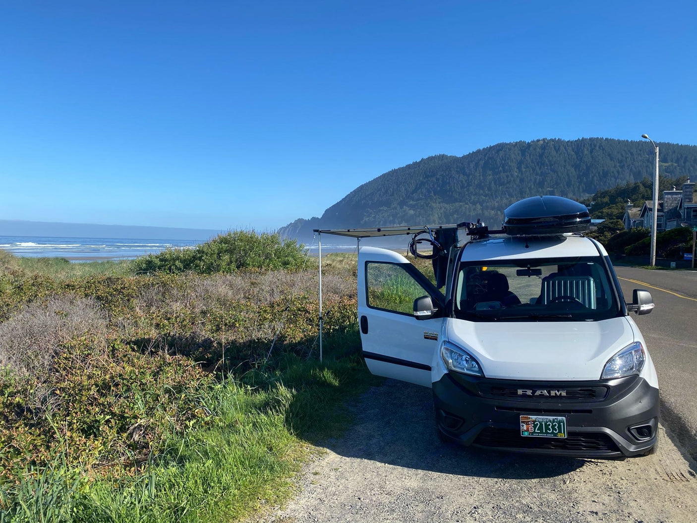 This RV rental, a white camper van, is parked overlooking a beautiful sunny ocean and coastal mountain view.