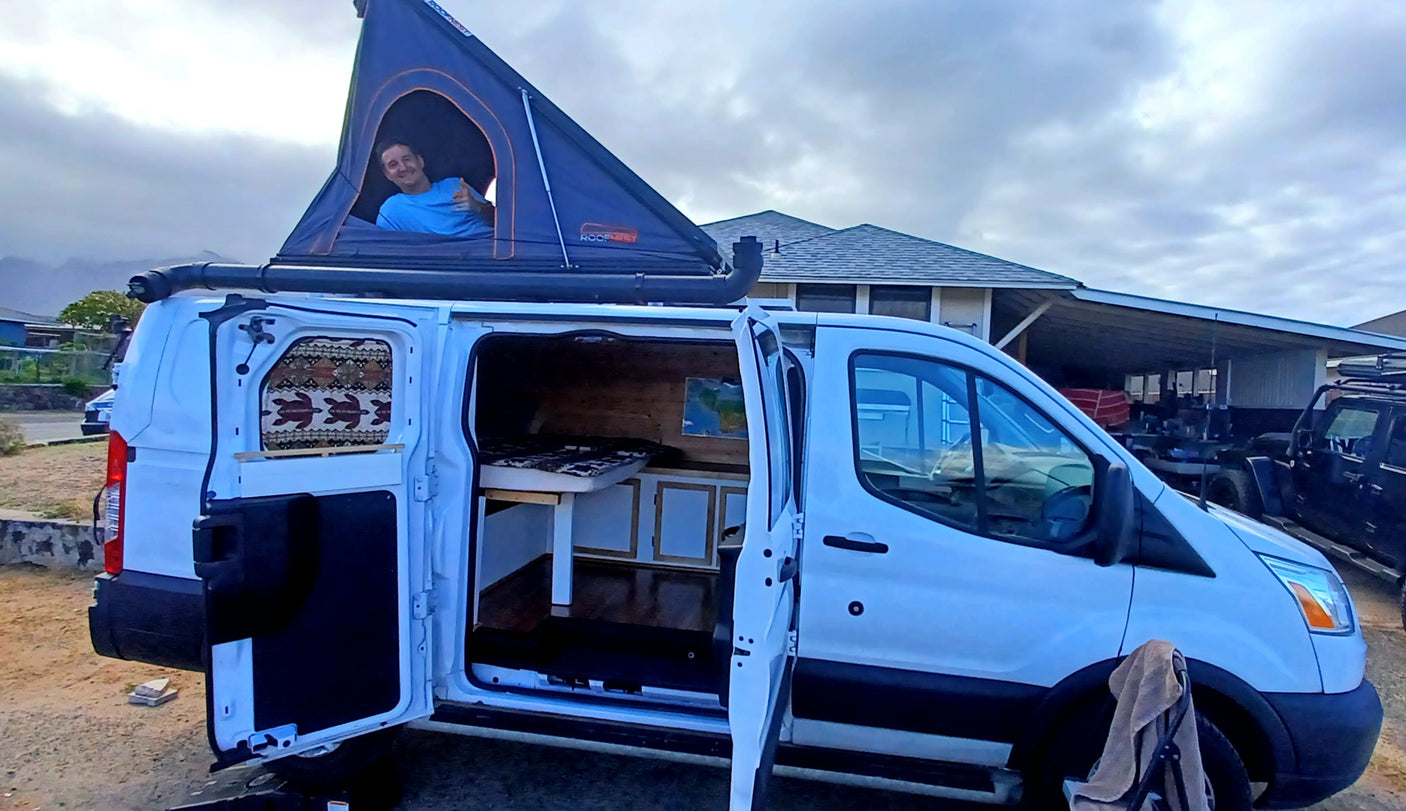 RV rental: A white Ford Transit camper van with an open rooftop tent, revealing a man smiling, ready for adventure.