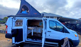 RV rental: A white Ford Transit camper van with an open rooftop tent, revealing a man smiling, ready for adventure.