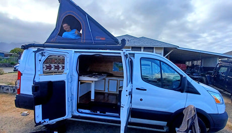 RV rental: A white Ford Transit camper van with an open rooftop tent, revealing a man smiling, ready for adventure.