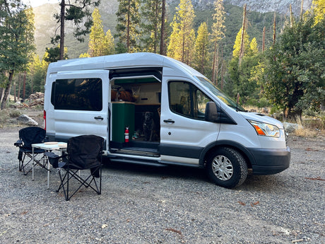 White rv rental camper van parked on gravel in a wooded mountain setting, open door reveals dog, with outdoor chairs & table.