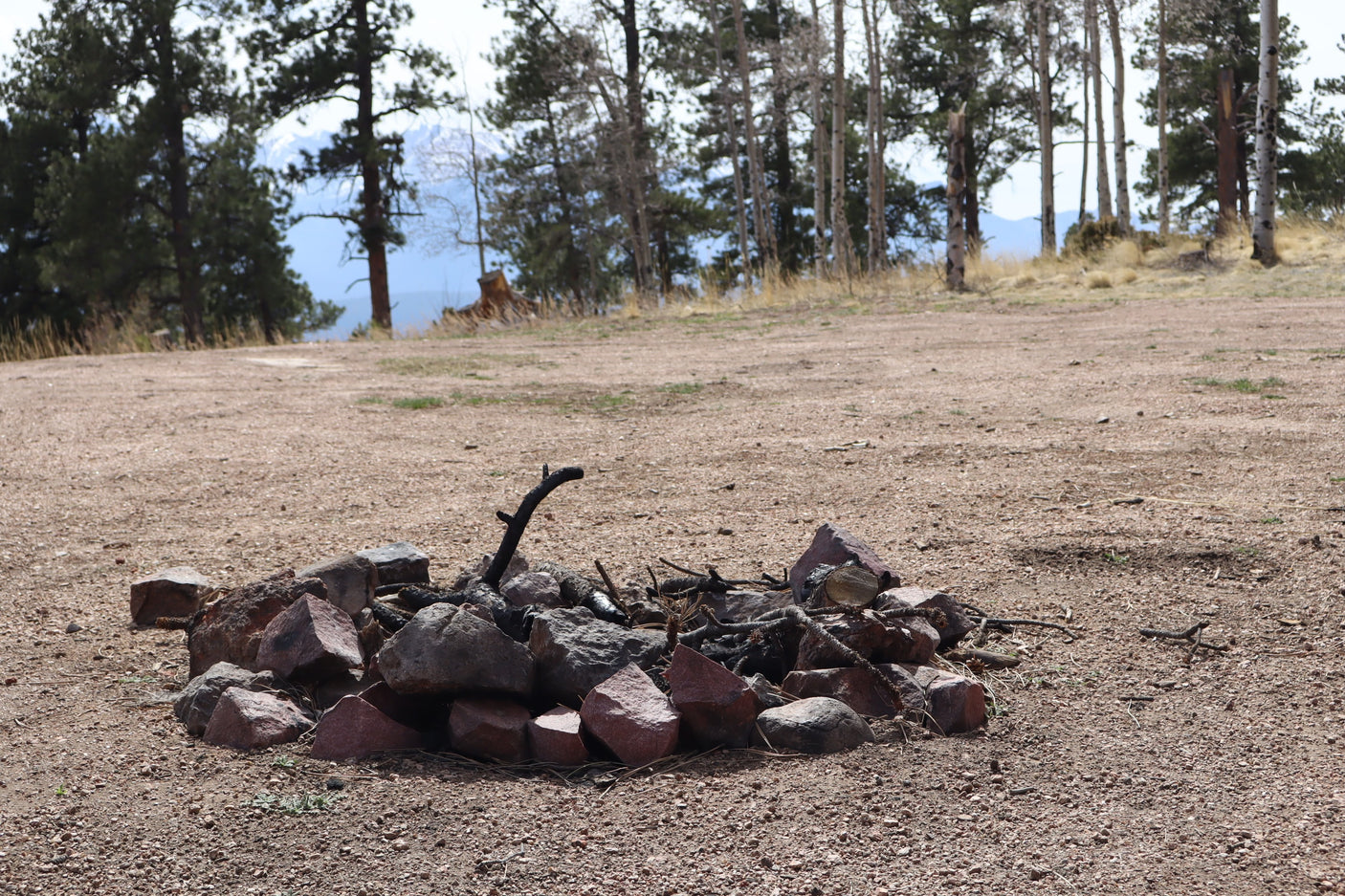 RV rental adventure awaits! A rock fire pit in a gravel clearing, surrounded by a forest and distant mountains.