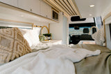 Modern RV rental interior with a cozy bed, compact kitchen, and overhead storage. Enjoy scenic views while traveling.