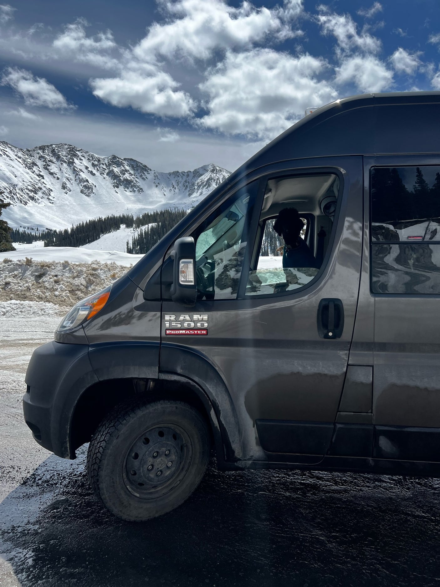 Gray Ram ProMaster RV rental parked amidst snowy mountains. A dog looks out the driver's window, enjoying the winter mountain view.