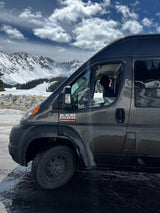 Gray Ram ProMaster RV rental parked amidst snowy mountains. A dog looks out the driver's window, enjoying the winter mountain view.