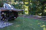 RV rental camper, with rooftop tent, solar panels, & awning, set up in a grassy yard next to trees and a fence.