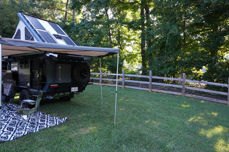 RV rental camper, with rooftop tent, solar panels, & awning, set up in a grassy yard next to trees and a fence.