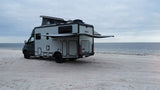 This light grey rv rental, a Winnebago EKKO with its pop-top extended and awning out, is parked on a sandy beach next to the ocean.