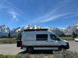 White Storyteller Overland RV rental van parked before the majestic, snow-capped Grand Teton mountains under a blue sky.
