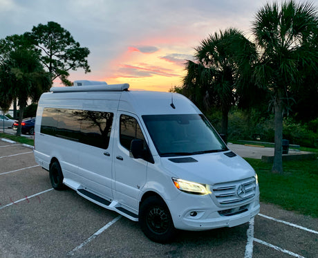 White Mercedes Sprinter RV rental parked at dusk. Features side steps & roof unit, with a colorful sunset sky.