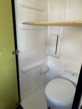 Compact white wet bath in an RV rental, featuring a toilet, storage access panel, and wooden shelf. Green door visible.