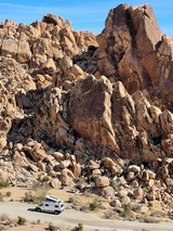 White RV rental parked in a rugged desert landscape among massive rock formations under a clear blue sky.