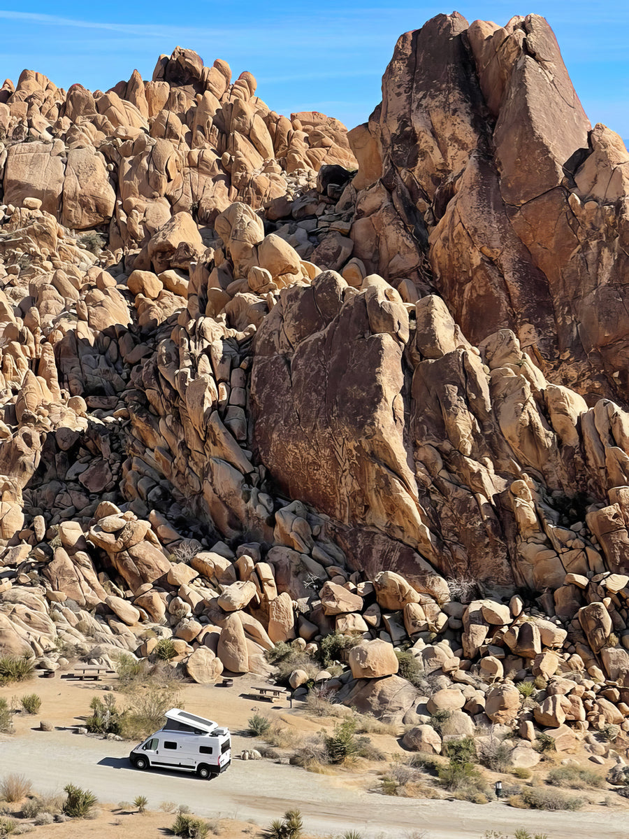 White RV rental parked in a rugged desert landscape among massive rock formations under a clear blue sky.