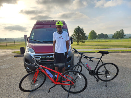 A man in a yellow helmet and white shirt stands with two bikes (red & black) by a red RV rental van in a park.
