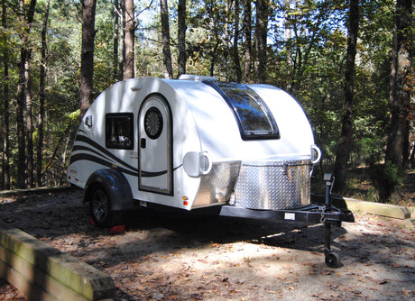 A white teardrop RV rental parked on a leaf-covered campsite, nestled among tall trees in a sunny forest.
