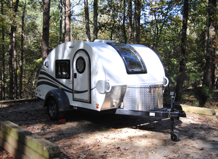A white teardrop RV rental parked on a leaf-covered campsite, nestled among tall trees in a sunny forest.