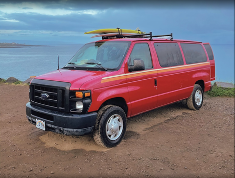 This red Ford van, with a surfboard on its roof, is parked seaside—perfect for an RV rental or coastal adventure.
