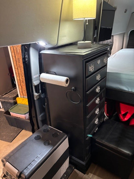 This RV rental features efficient van life design: a tall dresser with a lamp, TV, cooler, and ottoman provides compact storage.