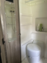 An RV rental's compact wet bath showing a white toilet, shower area with toiletries, and a mirror on the wooden door.
