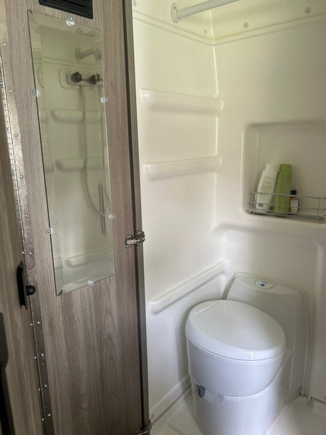 An RV rental's compact wet bath showing a white toilet, shower area with toiletries, and a mirror on the wooden door.