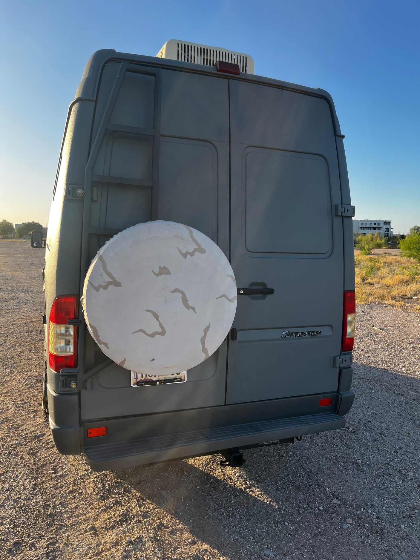 Rear view of a gray Mercedes Sprinter rv rental camper van, featuring a spare tire, ladder, roof AC, and trailer hitch.