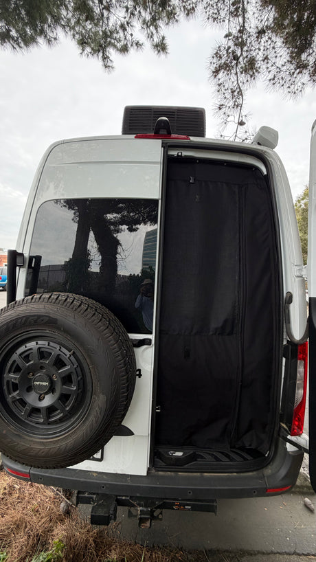 Rear of a white rv rental van with open door revealing a black privacy screen. Spare tire on left door. Tree reflections in window.