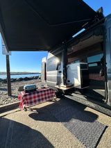 An RV rental parked on a sandy shore, ready for outdoor cooking with its awning extended and door open.