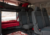 Inside an RV rental van: bunk beds with red and black bedding, rear passenger seats, and an outdoor view, ready for travel.
