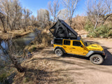 Yellow Jeep Wrangler with a rooftop tent pitched by a river, ideal for overland camping adventures. A unique rv rental alternative.