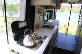 Enjoy your rv rental! Compact camper van kitchen with a stovetop, sink, and kettle, overlooking a serene lake.