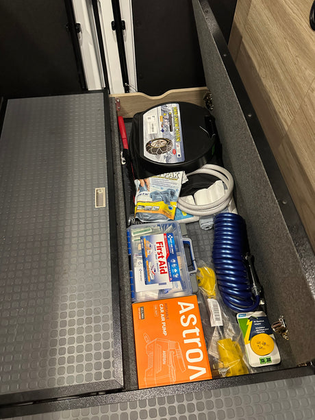 An organized RV rental compartment holds snow chains, a first aid kit, a car air pump, and an RV electrical adapter.