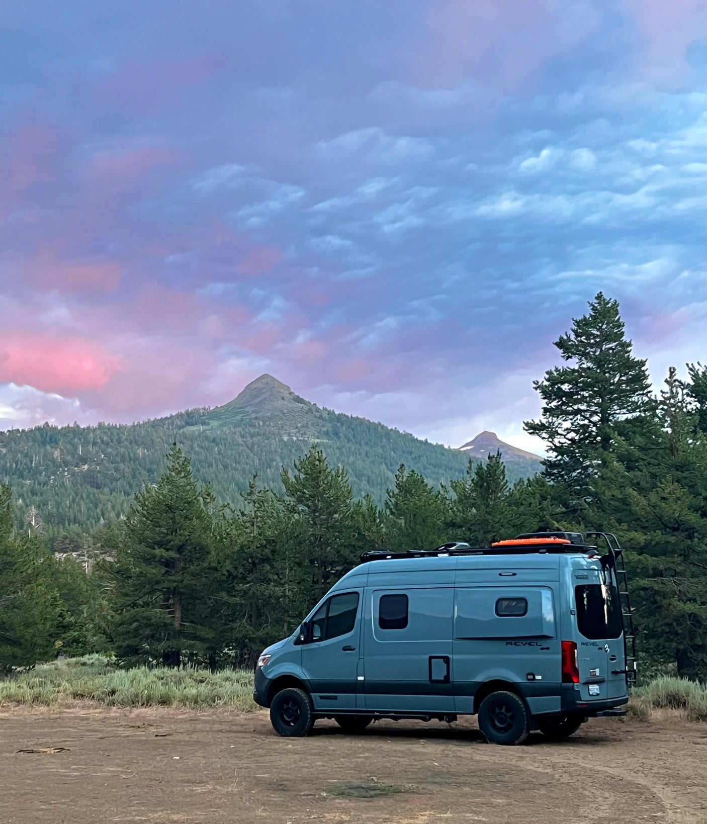 Grey Revel RV rental in a mountain forest. Vibrant purple and pink sky over peaks. Ideal for outdoor adventures.