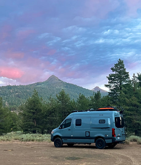 Grey Revel RV rental in a mountain forest. Vibrant purple and pink sky over peaks. Ideal for outdoor adventures.
