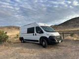 White RV rental van parked on a dirt road in a dry, hilly landscape under cloudy skies. Perfect for a camper van road trip.