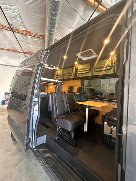 Step inside this custom camper van: black seats, wood table, fridge, and overhead storage. Ideal for your next RV rental adventure.