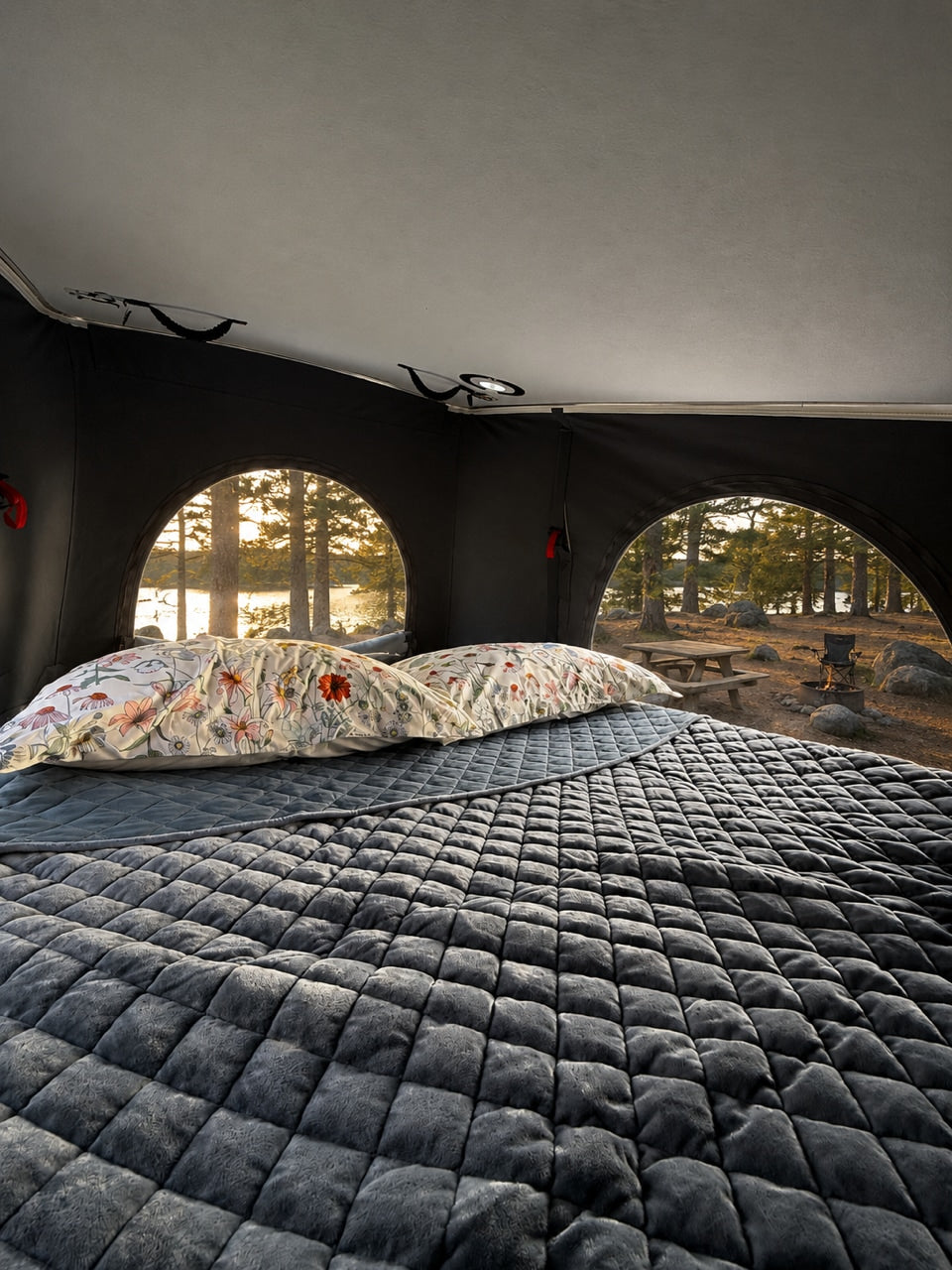 Cozy rv rental interior with a plush, quilted bed & floral pillows. Arched windows reveal a beautiful lake & forest view.