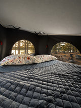 Cozy rv rental interior with a plush, quilted bed & floral pillows. Arched windows reveal a beautiful lake & forest view.
