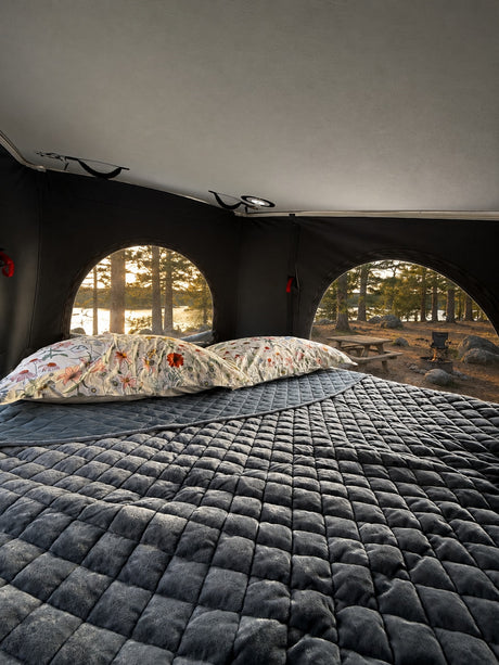 Cozy rv rental interior with a plush, quilted bed & floral pillows. Arched windows reveal a beautiful lake & forest view.