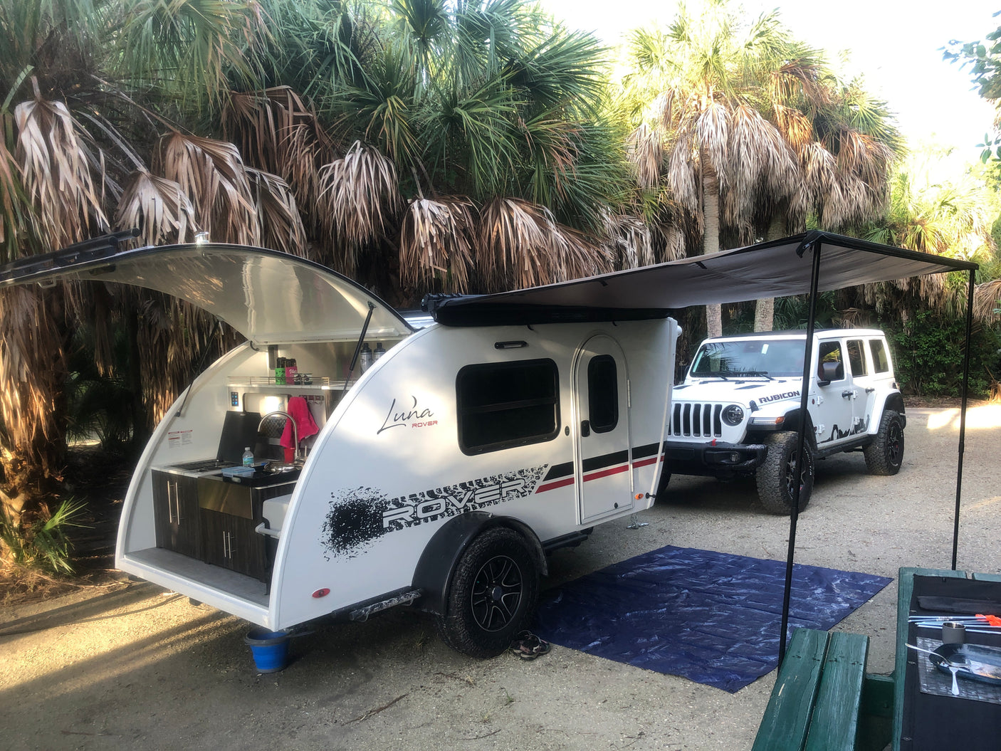 RV rental: White teardrop trailer with outdoor kitchen & awning, towed by a Jeep at a palm-lined campsite. Perfect for camping!