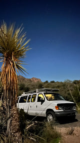 A white camper van, an ideal RV rental, illuminated under a clear, star-filled desert night sky with a tall yucca.