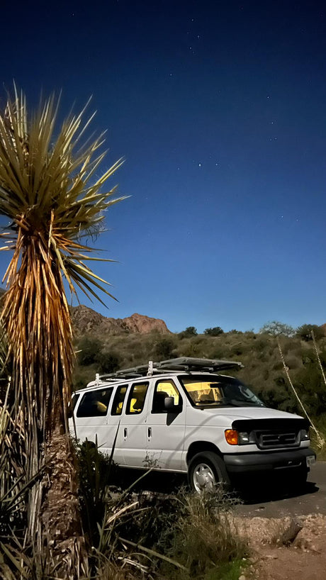 A white camper van, an ideal RV rental, illuminated under a clear, star-filled desert night sky with a tall yucca.