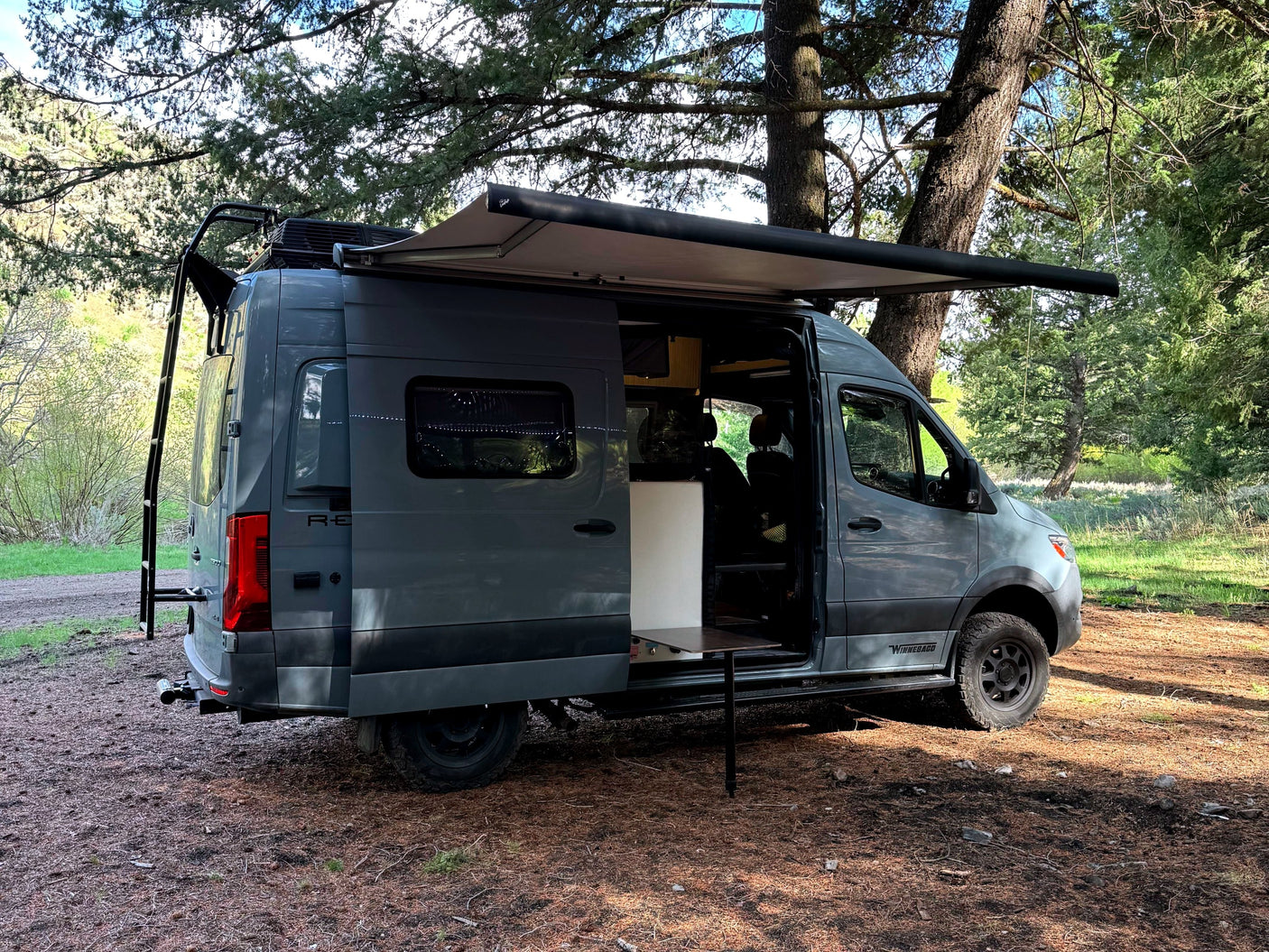 Gray camper van for rv rental parked in a forest with an extended awning and open side door, showcasing outdoor living.