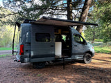 Gray camper van for rv rental parked in a forest with an extended awning and open side door, showcasing outdoor living.