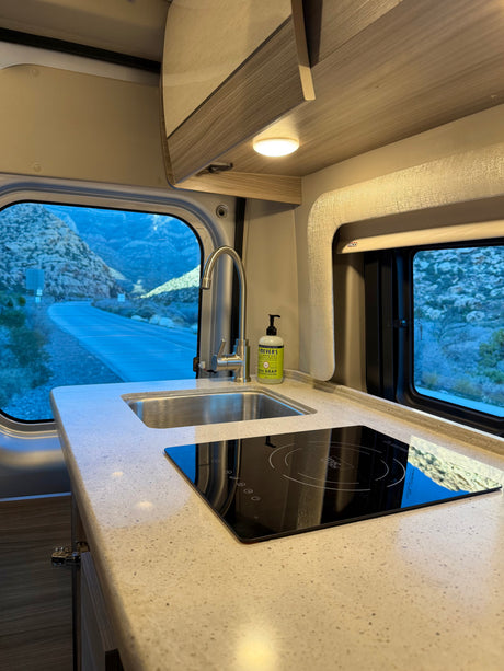 RV rental kitchen: modern sink, induction cooktop, and stunning mountain road views from a camper van window.