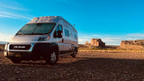 A white camper van, perfect for RV rental adventures, parked on a gravel road in a vast desert landscape with large rock formations under a clear blue sky.