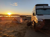 Stunning sunset camping scene with an RV rental, tent, and chairs by the water. Perfect outdoor adventure.