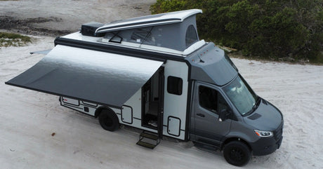 A grey and white rv rental camper van with an extended awning and raised pop-top roof, parked on sand.